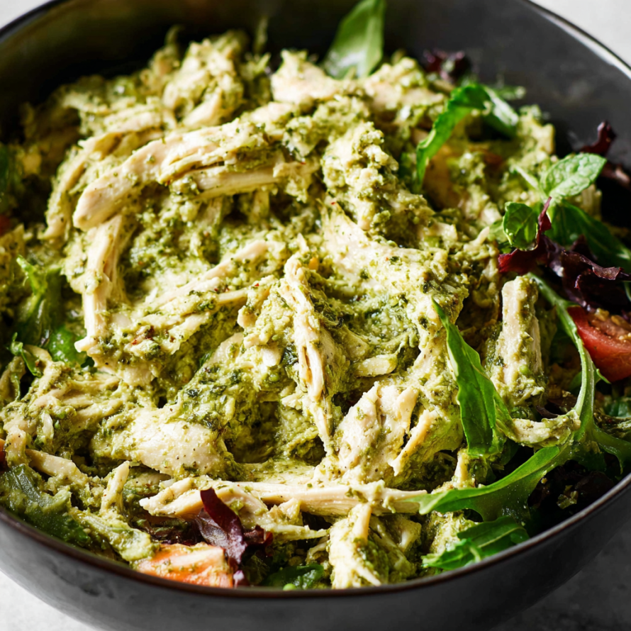 A bowl of chicken salad with pesto sauce, tomatoes, and greens.