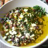A white bowl filled with a Greek-inspired bread dipping oil, containing olives, cheese, and herbs.