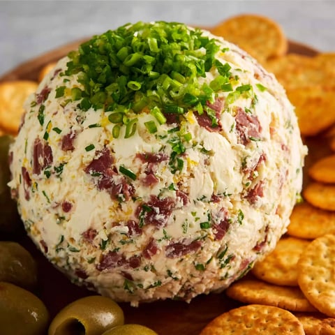 A cheese ball with a green topping sits on a wooden tray, accompanied by crackers and olives.